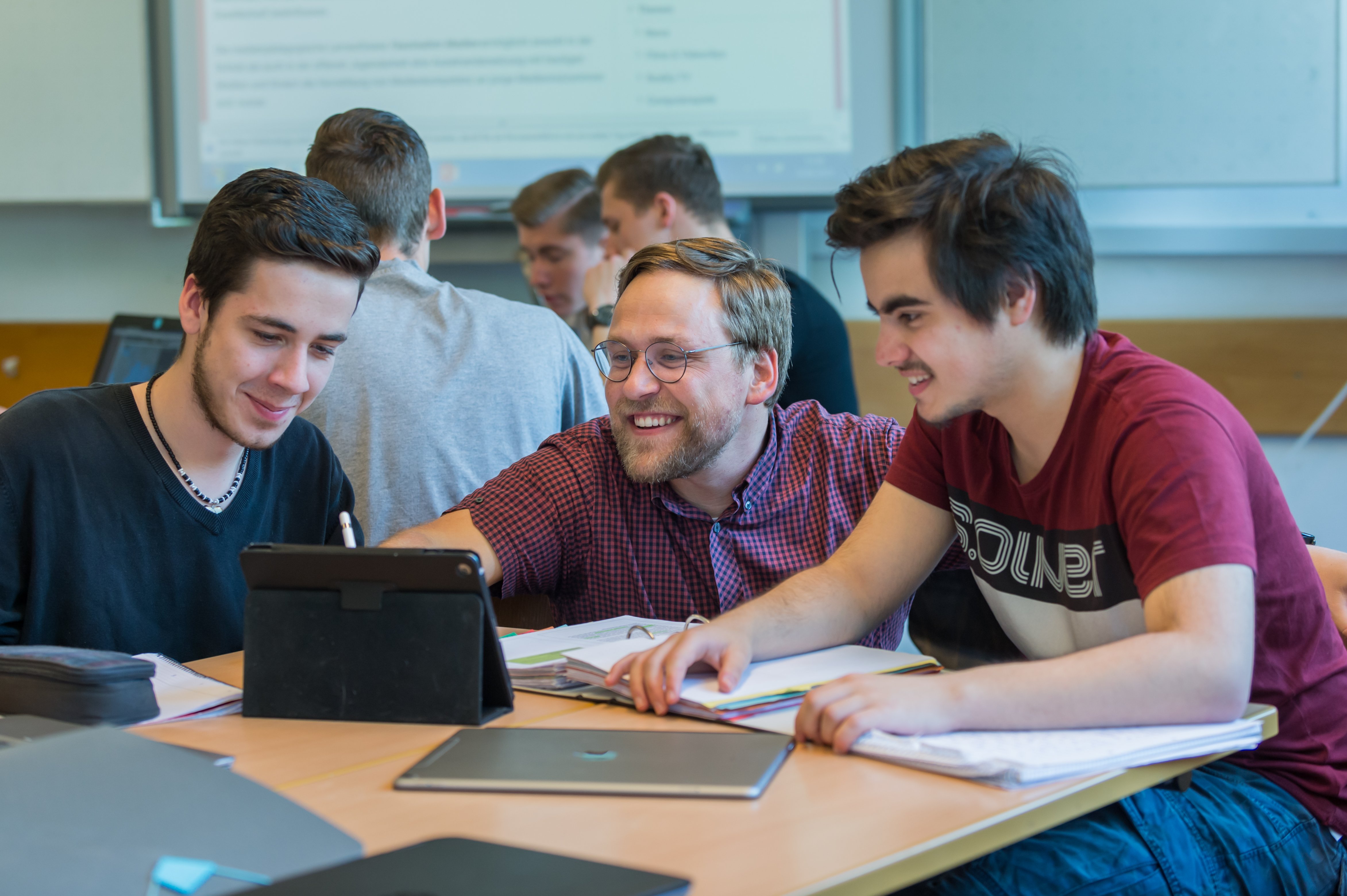 Ein Lehrer im Gespräch mit zwei Schülern, die an Tablets arbeiten.