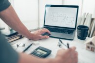 Zu sehen ist der Arbeitsplatz eines Bauzeichner mit Laptop, Taschenrechner, Plänen und anderem.