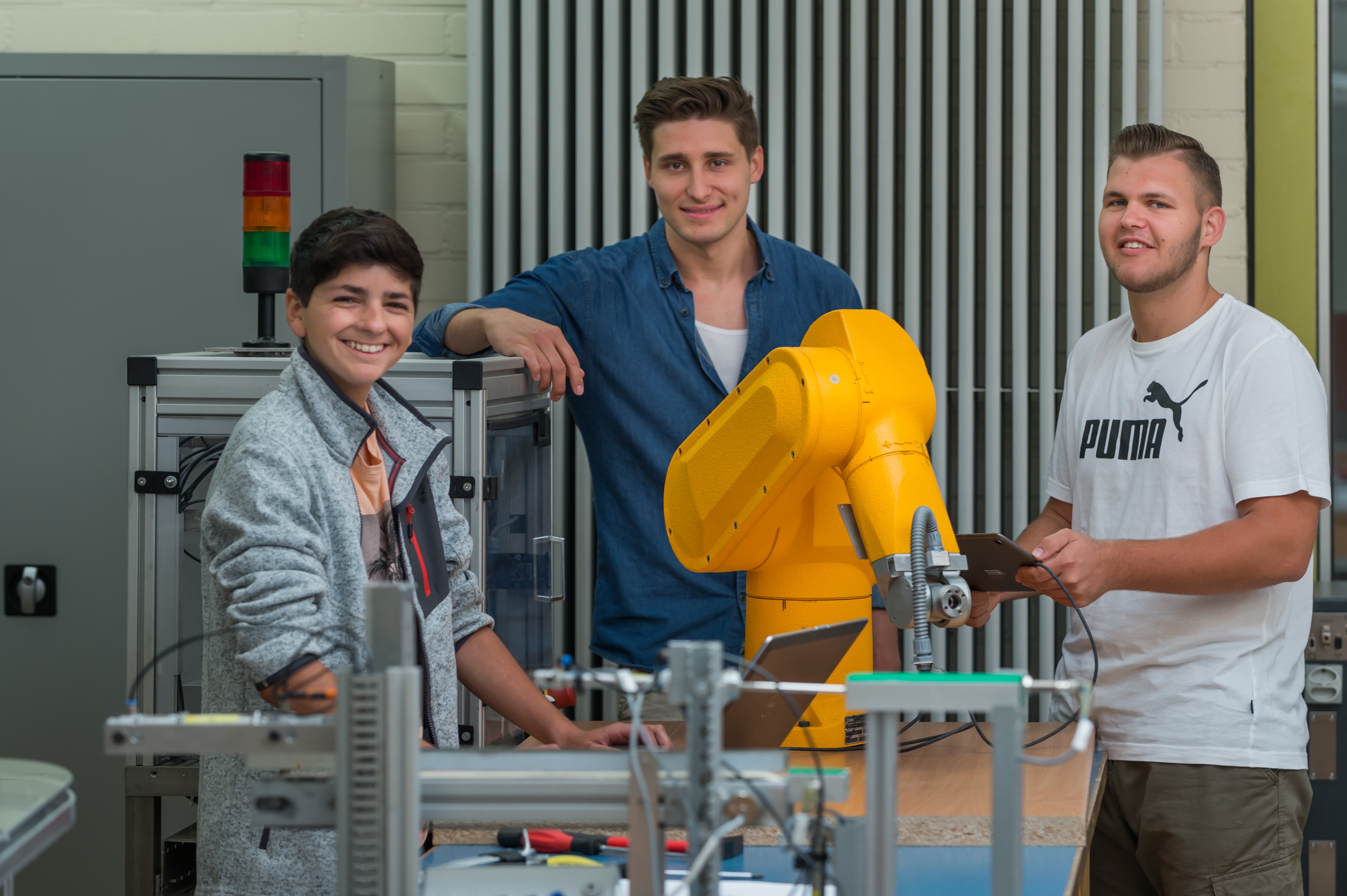 Drei Schüler aus dem Bereich Mechatronik stehen an einer Maschine und lächeln