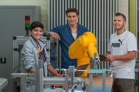 Drei Schüler aus dem Bereich Mechatronik stehen an einer Maschine und lächeln