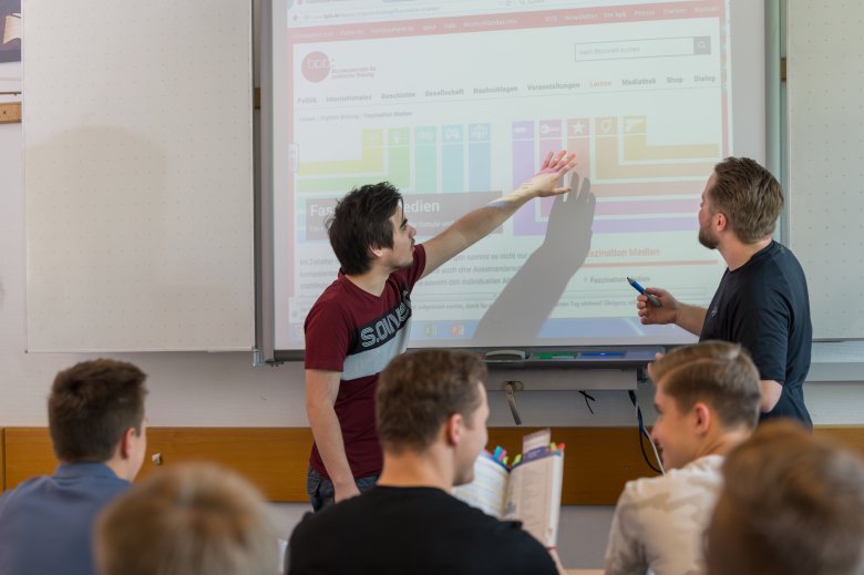 Schüler und Lehrer stehen vor der Projektion einer Präsentation und der Schüler zeigt auf einen Inhalt dessen