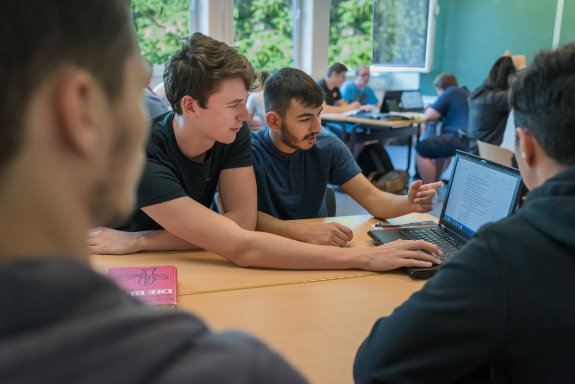 Zwei Schüler im Fokus sitzen im Klassenraum mit ihren Mitschülern; die beiden schauen etwas am Laptop