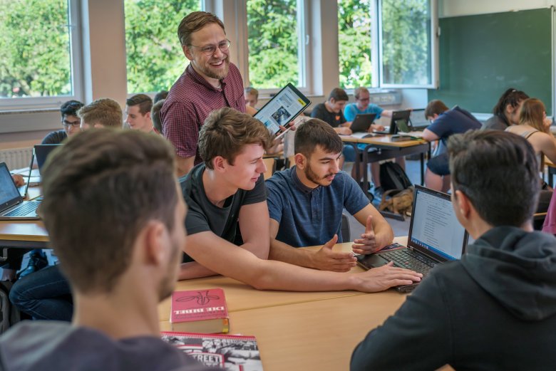 Ein Lehrer mit Tablet in der Hand im Gespräch mit Lernenden. Alle sind gut gelaunt.