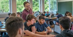 Ein Lehrer mit Tablet in der Hand im Gespräch mit Lernenden. Alle sind gut gelaunt.
