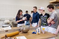 Eine Lehrerin liest ihren Schülern ein Rezept vor. Sie befinden sich in einer großen Backstube.