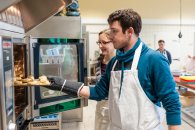 Eine Lehrerin und ein Schüler schieben ein Blech mit Backwaren in den Ofen.