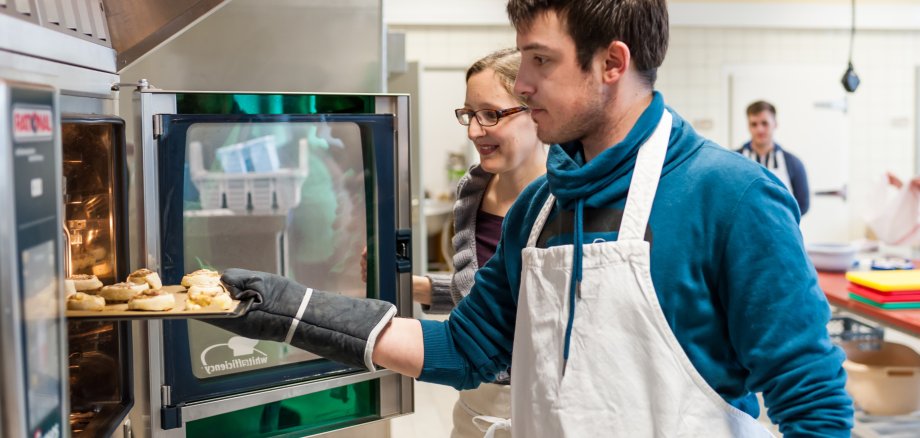 Schüler in Schürze Schiebt ein Bleck mit Pizzaschnecken in den Ofen. Dahinter steht eine Lehrerin und beobachtet den Vorgang