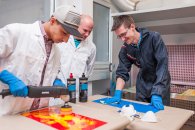 Drei Schüler in Schutzkleidung schleifen Metallplatten ab, um diese zu lackieren.