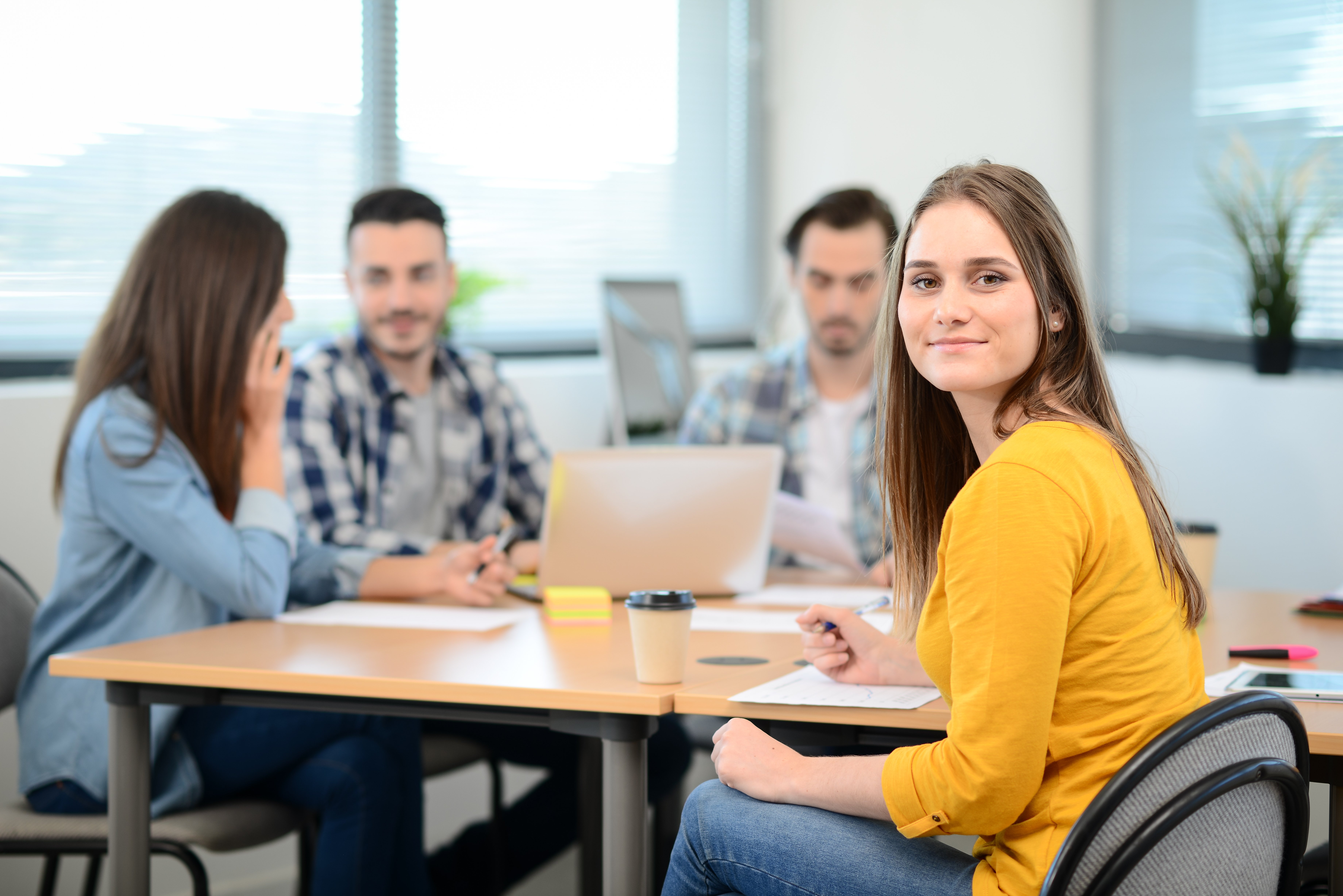Portrait einer Frau an einem Besprechungstisch, im Hintergrund sitzen weitere Personen und arbeiten an etwas