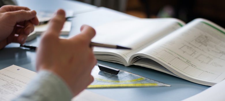Ein Tisch, auf dem ein Geodreieck und diverse Unterrichtsmaterialien in Form von Arbeits- und Notizheften angeordnet, sowie ein Arm und eine Hand, die auf dem Tisch abgelegt sind.